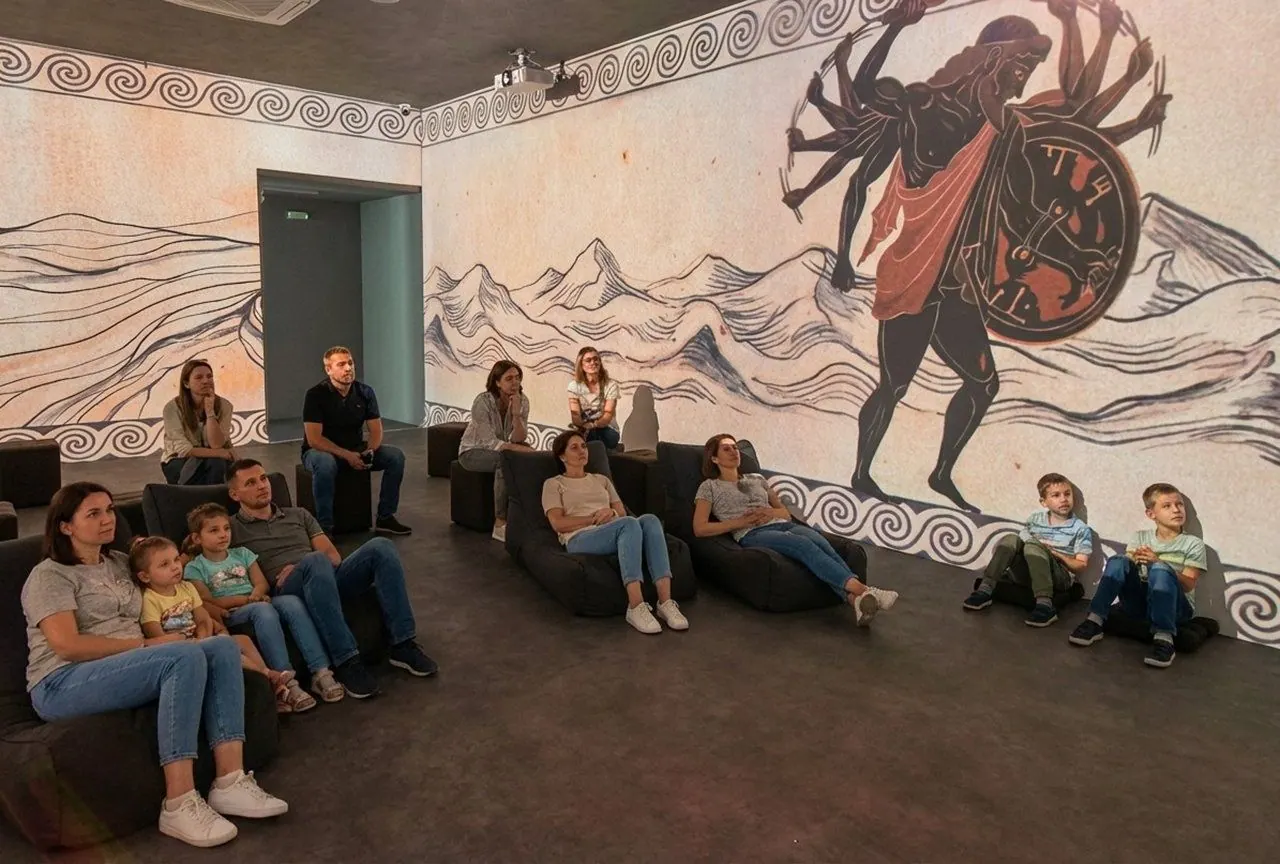 Παιδιά και γονείς κάθονται στην immersive αίθουσα, ενώ στον τοίχο προβάλλεται μια μορφή από τη μυθολογία πάνω από τα βουνά.